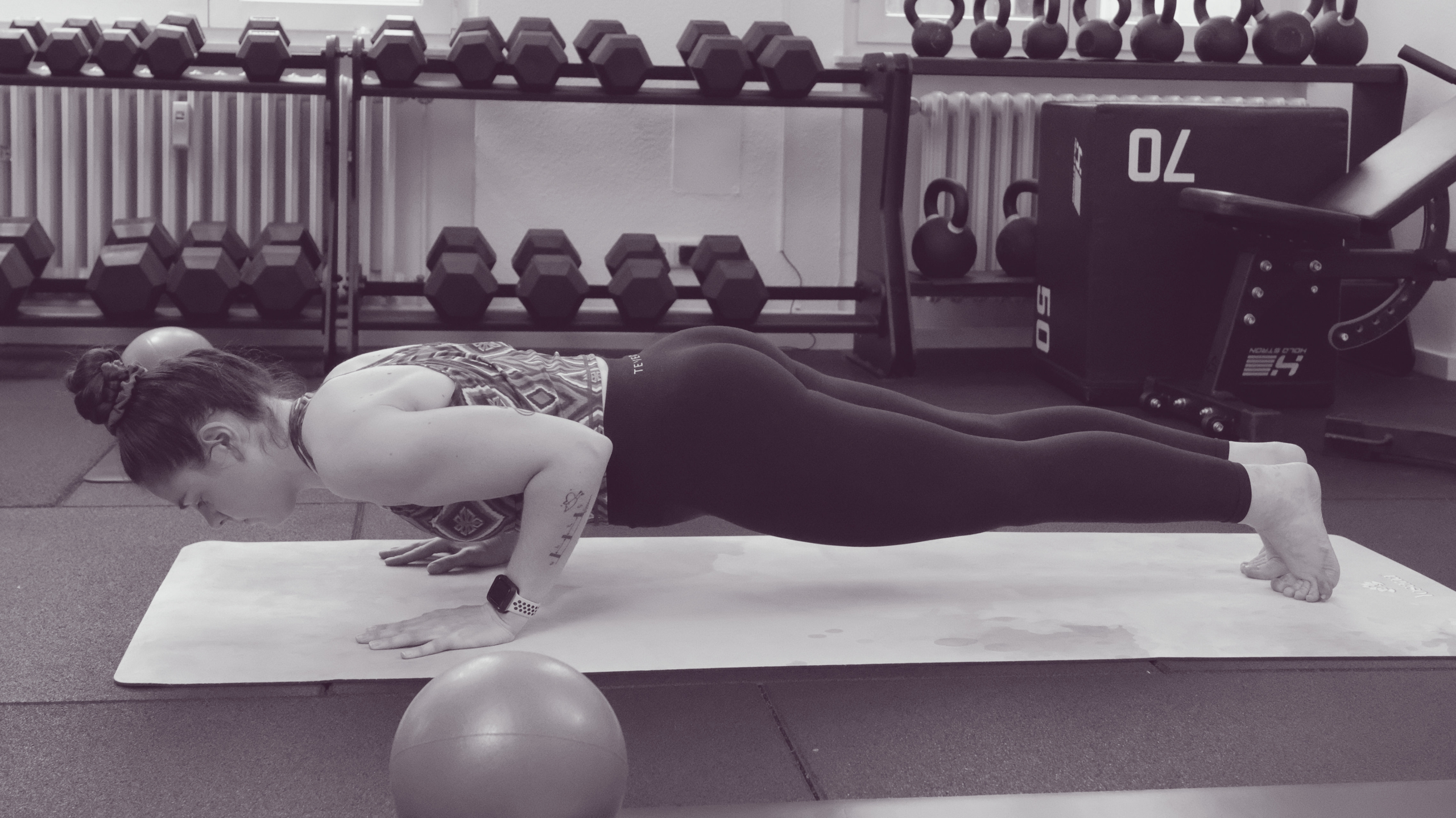 Frau hält im Fitnessstudio eine Plank Position vor einem Regal mit Kurzhanteln